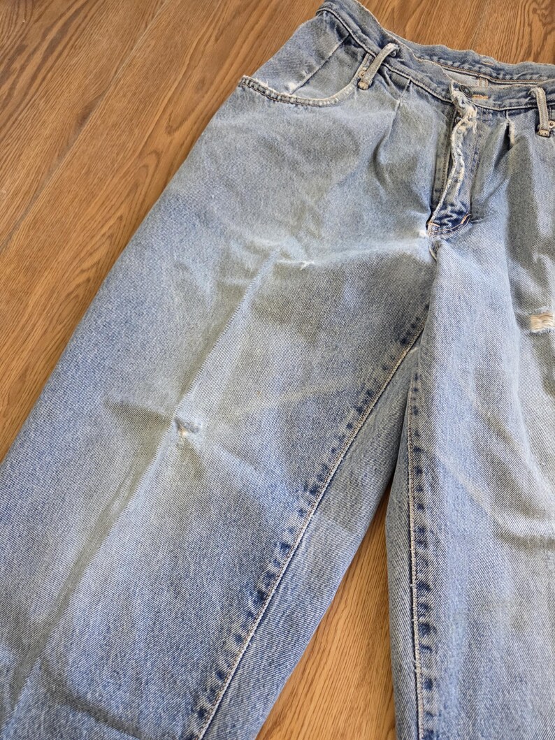 May include: Light blue denim jeans with a faded wash and distressed details. The jeans have a classic five-pocket design, a zipper fly, and a button closure. The fabric shows signs of wear and tear, adding to the vintage aesthetic.