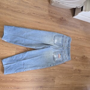 May include: Light blue denim jeans with distressed details, including rips on the front and back pockets. The back features a logo patch. The jeans are laid flat on a wooden surface.