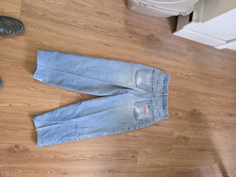 May include: Light blue denim jeans with distressed details, including rips on the front and back pockets. The back features a logo patch. The jeans are laid flat on a wooden surface.