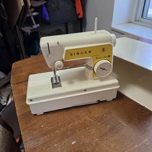 Peut inclure: Machine à coudre Singer Touch & Sew vintage, crème et jaune, posée sur une table en bois. La machine comprend une aiguille, une bobine de fil et un cadran. Le logo Singer est visible à l'avant.