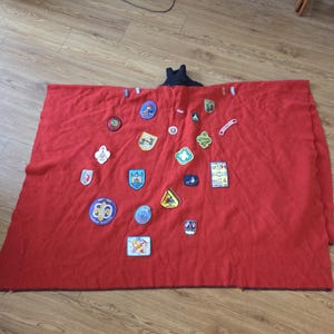 May include: A red blanket with various colorful patches sewn on. The patches feature different shapes and designs, including badges with text and images. The blanket is spread out on a wooden floor.