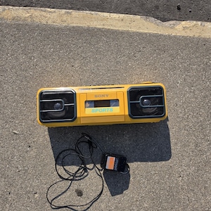 May include: A yellow Sony Sports cassette player featuring two black speaker grilles on the sides and a central cassette compartment with the text SONY and SPORTS printed in blue. A black power adapter with a long cord is attached to the device.