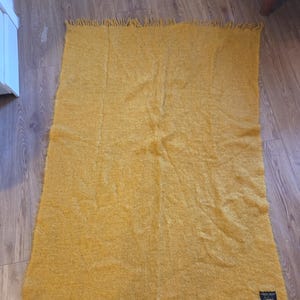 May include: Mustard yellow rectangular throw blanket with fringed edges. The blanket has a textured, woven appearance. A small, dark square label is attached to the blanket. The blanket is lying on a wooden floor.