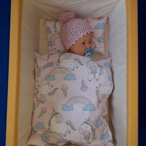 Könnte beinhalten: Eine Puppe mit einem rosa gehäkelten Hut und einem Schnuller, die in einem kleinen gelb-weißen Kinderbett liegt. Die Puppe liegt auf einem Kissen und unter einer Decke mit einem rosa Einhorn- und Regenbogenmuster.