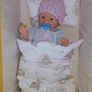 Può includere: Una bambola con un cappello rosa all'uncinetto con un pompon, un ciuccio blu e una tutina azzurra. La bambola è adagiata in una culla bianca con un cuscino e una coperta con un motivo ripetuto di orsi, nuvole e stelle.