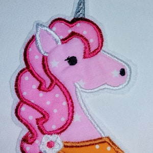 Puede incluir: Aplique de unicornio rosa con lunares blancos, un cuerno plateado y un cuello naranja y blanco con lunares.