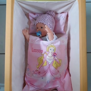 Könnte beinhalten: Ein rosa-weißes Puppenbett mit einer Puppe, die einen lila Hut und einen Schnuller trägt. Das Bett hat ein rosa-weißes Bettwäsche-Set mit einem Prinzessinnen-Design und dem Namen "Emma" darauf gedruckt.
