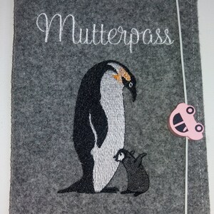 Könnte beinhalten: Grauer Filzbezug mit dem in weiß gestickten Wort "Mutterpass". Der Bezug zeigt eine gestickte Abbildung von zwei Pinguinen, einem großen und einem kleinen, wobei der größere Pinguin aufrecht steht und der kleinere Pinguin auf seinen Füßen mit ausgebreiteten Flügeln steht. Ein rosa Plastikclip in Form eines Autos ist am Bezug befestigt.