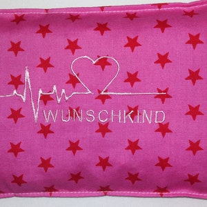 Puede incluir: Almohada de tela rosa con estrellas rojas y bordado blanco que dice "Wunschkind" con un corazón y una línea de latido del corazón.