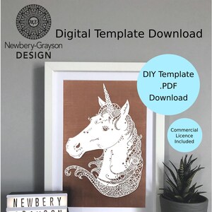 Op de afbeelding: Een witte papieren uitsnijding van een eenhoornhoofd met ingewikkelde details, ingelijst in een wit frame met een bruine achtergrond. Het frame staat op een plank met een kleine potplant en een lichtbak met de tekst "NEWBERY GRAYSON DESIGN". De tekst "DIY Template .PDF Download" staat in een blauwe cirkel boven het frame. De tekst "Commercial Licence Included" staat onder het frame.