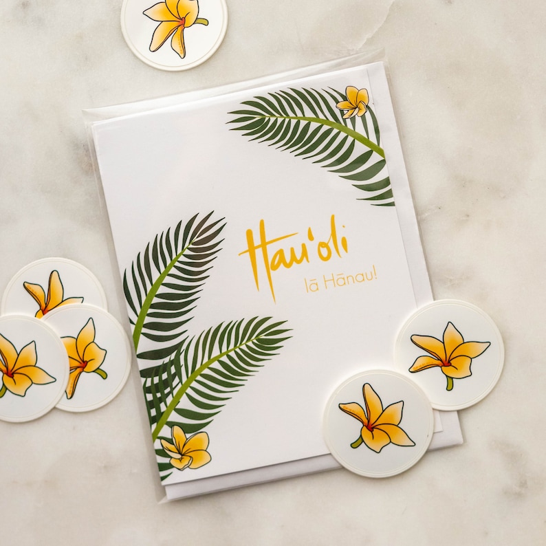 May include: Greeting card with tropical theme.  Features yellow plumeria flowers and dark green palm fronds.  Text on card says 'Hau'oli La Hanau!'  Comes with several small, round stickers featuring the same yellow plumeria flower design.  The card and stickers are packaged together.