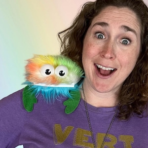 May include: A woman wearing a purple shirt with the word "VERT" printed on it. She is smiling and has a rainbow-colored fuzzy creature on her shoulder. The creature has large eyes and green claws.