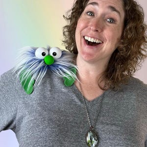 May include: A plush shoulder puppet featuring long, shaggy blue and white faux fur, two large white plastic eyes, a bright green round nose, and two green felt leaf-like appendages extending from the sides of the creature's body.