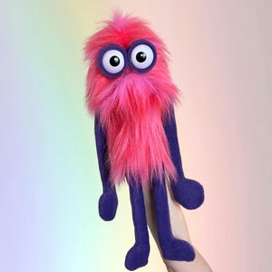 May include: A whimsical, handmade puppet with a vibrant pink, shaggy fur body and a matching tuft on top. It has large, googly eyes with purple rims and long, purple felt arms and legs. The puppet is designed to be held.