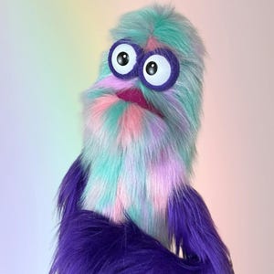 May include: A plush puppet featuring long, shaggy faux fur in shades of teal, pink, and lavender. The creature has two large, round white eyes with black pupils, each surrounded by a thick purple felt ring, and a small, dark pink felt mouth.
