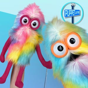 May include: Two colorful, fuzzy puppets with large, expressive eyes. One puppet is primarily pink, yellow, and blue, while the other is blue, yellow, and white. Both have pink arms and legs. The sign in the background reads "Puppet Layne."