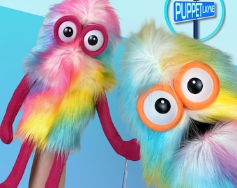 May include: Two colorful, fuzzy puppets with large, expressive eyes. One puppet is primarily pink, yellow, and blue, while the other is blue, yellow, and white. Both have pink arms and legs. The sign in the background reads "Puppet Layne."