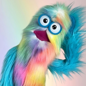 May include: A hand puppet covered in long, shaggy faux fur featuring a vibrant rainbow gradient of blue, green, yellow, and pink. The puppet has two large, round white eyes with black pupils set in blue rings and a deep purple, open mouth interior.