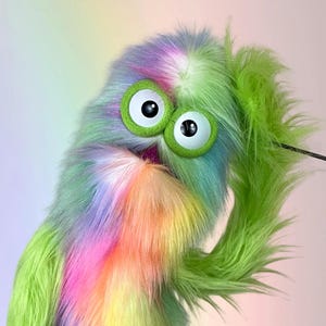 May include: A colorful, furry puppet with large, expressive eyes. The puppet's fur is a mix of green, purple, pink, and yellow. It has large, round, green-rimmed eyes with black pupils. The puppet appears to be looking at the viewer.