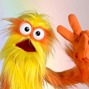 May include: A fuzzy puppet with long yellow and orange fur, featuring two large white eyes with black pupils set in orange circular frames. The puppet has a dark red open mouth and an orange fleece-covered hand raised in a peace sign gesture.