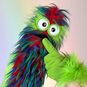 May include: A fuzzy puppet with long, shaggy fur in vibrant shades of green, blue, and red. It features two large, round white eyes with black pupils and a wide, dark mouth. A bright green, fleece-covered arm is raised, showing a thumbs-up gesture.