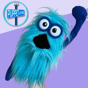May include: A blue puppet with large white-and-black eyes, a black mouth, and shaggy blue fur. The puppet has a raised arm and is set against a purple background. A sign that says "Puppet Layne" is in the upper left corner.