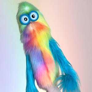 May include: A tall, plush puppet covered in long, shaggy faux fur featuring a vibrant rainbow gradient of green, yellow, orange, pink, and blue. It has two large, round, white eyes with black pupils, each framed by a thick, bright blue circular border.