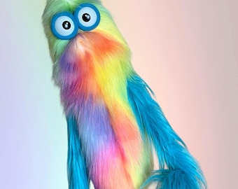 May include: A whimsical, colorful puppet with a fluffy, rainbow-colored body and large, cartoonish blue eyes. The puppet has a green head, a yellow and pink chest, and blue feathery accents.