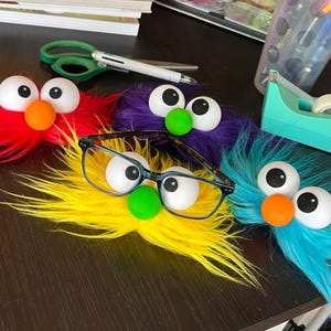 May include: Four fuzzy monster-themed decorations with large white googly eyes and colorful noses. One red, one purple, one yellow, and one blue. A pair of dark-rimmed eyeglasses rests on the yellow decoration, which has a bright green nose.