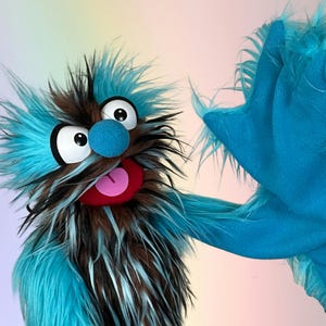 May include: A whimsical puppet with a blue, brown, and white furry head and a large, open mouth with a pink tongue. The puppet has large, round eyes and a blue nose. A blue furry arm is visible on the right side. The puppet is set against a soft, colorful background.
