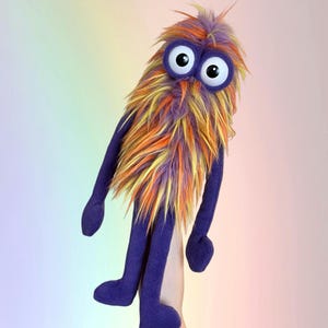May include: A whimsical, handmade puppet with a fuzzy, colorful body. The puppet features large, googly eyes and a vibrant mix of orange, yellow, and purple fur. The arms and legs are a solid purple color, creating a playful and unique design.