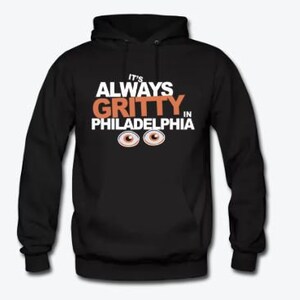 Op de afbeelding: Zwart hoodie met witte tekst die "It's Always Gritty in Philadelphia" zegt met twee oranje ogen onder de tekst.