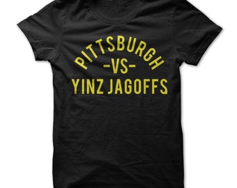 Op de afbeelding: Zwart T-shirt met een gele afbeelding met de tekst "Pittsburgh -VS- Yinz Jagoffs"