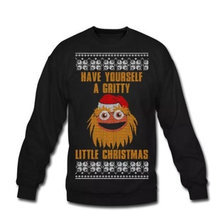 Op de afbeelding: Zwarte kersttrui met een bruin en oranje pluche monster dat een kerstmanmuts draagt. De trui heeft de tekst "Have Yourself a Gritty Little Christmas".