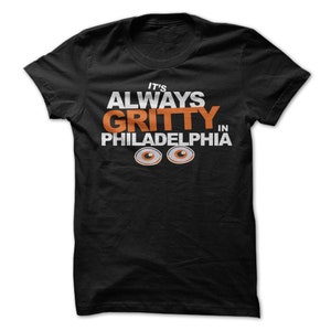 Op de afbeelding: Zwart T-shirt met witte tekst die "It's Always Gritty in Philadelphia" luidt met twee oranje ogen onder de tekst.