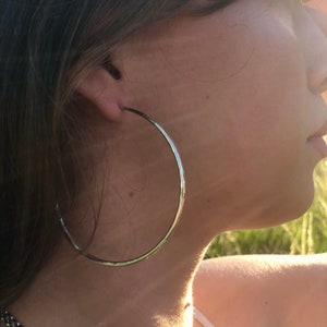 May include: Silver hoop earrings with a hammered finish. The earrings are large and circular, and they are worn by a person with long brown hair.