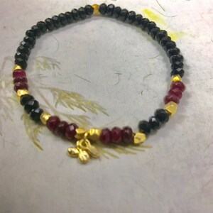 May include: A black and red beaded bracelet with a gold charm. The bracelet is made with small, round beads and has a delicate, feminine look.