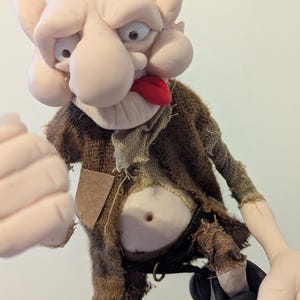 May include: A handcrafted doll with a disgruntled expression. The doll features a pale face, a red tongue, and is dressed in a ragged brown outfit. The doll's arms are extended, and it seems to be made of clay or a similar substance.