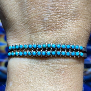 Puede incluir: Pulsera plateada con dos filas de vibrantes cabujones turquesas. La pulsera es de estilo brazalete, con las piedras turquesas muy juntas. Las piedras azul brillante contrastan con el metal plateado, creando una llamativa pieza de joyería.