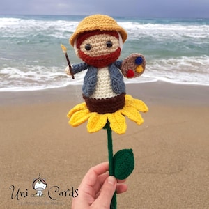 Könnte beinhalten: Eine gehäkelte Sonnenblume mit einer kleinen gehäkelten Figur eines Malers, der einen Pinsel und eine Palette hält. Die Figur trägt ein blaues Hemd, eine braune Hose und einen gelben Hut. Die Sonnenblume ist gelb mit grünen Blättern.