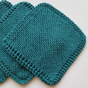 May include: Three teal knitted washcloths, square in shape. The washcloths are made from textured yarn and have a scalloped edge. They are stacked, with the top one partially covering the others. The background is white.