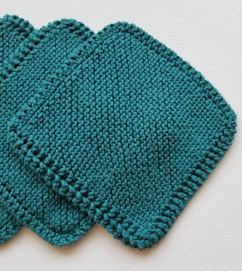 May include: Three teal knitted washcloths, square in shape. The washcloths are made from textured yarn and have a scalloped edge. They are stacked, with the top one partially covering the others. The background is white.
