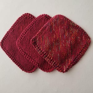 May include: Three handmade knitted washcloths in shades of burgundy. The washcloths are square with rounded corners and a textured knit pattern. One washcloth has a variegated yarn with colourful flecks.