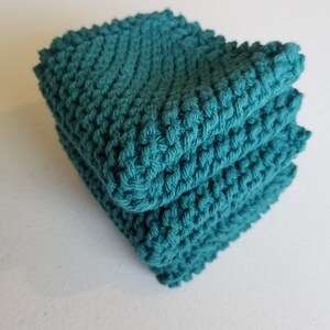 May include: A stack of three teal-coloured, hand-knitted washcloths. The washcloths have a textured stitch, creating a bumpy surface. The edges are slightly uneven, suggesting a handmade quality. The stack is neatly arranged, showcasing the colour and texture.