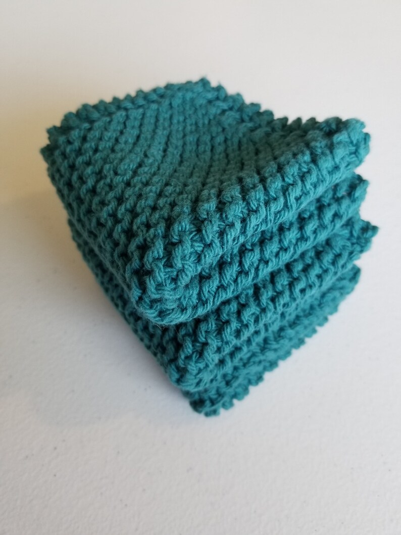 May include: A stack of three teal-coloured, hand-knitted washcloths. The washcloths have a textured stitch, creating a bumpy surface. The edges are slightly uneven, suggesting a handmade quality. The stack is neatly arranged, showcasing the colour and texture.