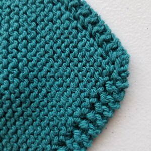 May include: Close-up of teal knitted fabric with a textured surface. The knitted material is a solid colour and appears to be made of yarn. The stitches are visible and create a pattern. The fabric is on a white surface.