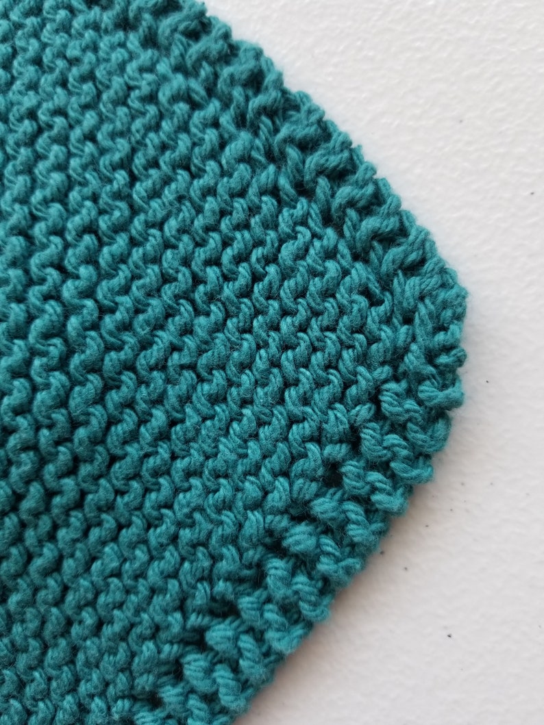 May include: Close-up of teal knitted fabric with a textured surface. The knitted material is a solid colour and appears to be made of yarn. The stitches are visible and create a pattern. The fabric is on a white surface.