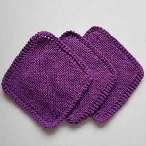 May include: Three square, hand-knitted washcloths in a vibrant purple hue. The washcloths are arranged in a slightly overlapping pattern, showcasing their textured surface and detailed edging. The soft, knitted texture suggests a gentle touch.