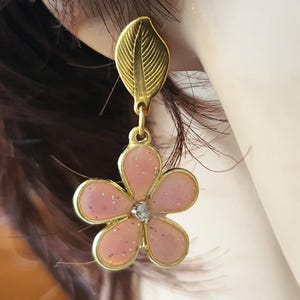 Op de afbeelding: Een goudkleurige oorbel met een gestructureerde, bladvormige knop verbonden aan een bungelende bedel in de vorm van een vijfbladige bloem. De bloem heeft een lichtroze, glinsterende emaille afwerking met een goudkleurige rand en een kristal.