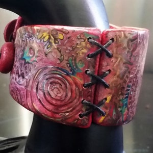 May include: A wide red and pink patterned bracelet with a black cord lacing up the side. The bracelet is made of polymer clay and has a unique, abstract design.
